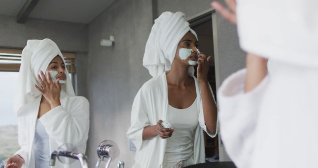 Women applying face masks while looking in bathroom mirror, both wearing towels on heads. Suitable for illustrating beauty routines, self-care tips, skincare product advertisements, home spas, and personal care promotions.