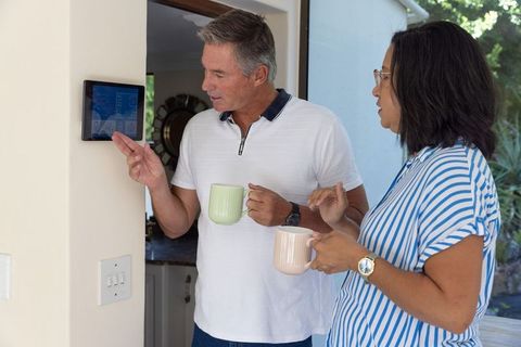 Mature diverse couple using smart home touchscreen while holding coffee mugs in entryway