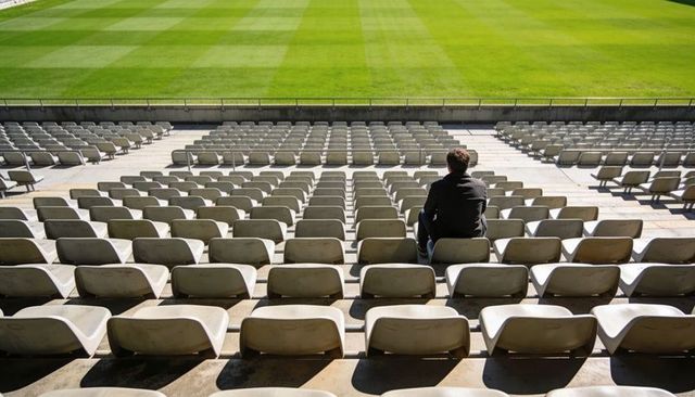 Lone spectator sitting on empty stadium bleachers facing sunlit striped grass field