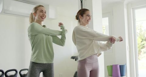 Two friends stretching together in bright home gym with yoga mats and kettlebells