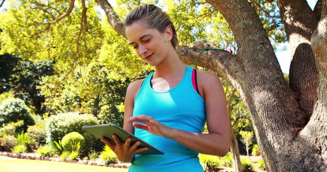 Young woman reading on a digital tablet while standing under a tree in a park. Great for concepts such as technology in nature, leisure activities, outdoor learning, and relaxation in natural settings.