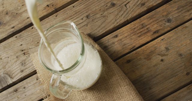 Image showing fresh milk being poured into a glass pitcher on a rustic wooden table with a burlap cloth. Perfect for use in food blogs, advertisements promoting dairy products, farmhouse decor items, natural living websites, or homestead lifestyle articles.