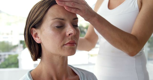 A Caucasian woman appears relaxed as another person places a hand on her forehead, in a gesture of healing or comfort, with copy space. Such an image could be used to depict wellness, alternative medicine, or a moment of empathy between individuals.