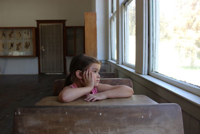 Young girl sitting at desk alone in empty classroom, gazing out of window. Captures feelings of solitude, nostalgia, and hope, often used for themes like education, childhood memories, daydreaming, loneliness. Excellent for articles, blogs, or covers focusing on children’s emotions or school life.