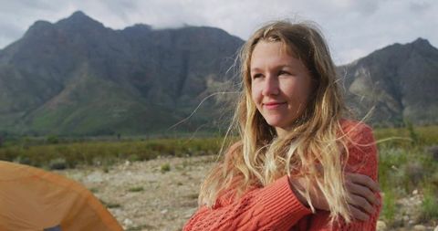 Young woman gazing toward mountain valley while camping, wearing orange sweater