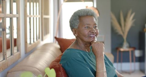 Thoughtful senior woman relaxing on leather sofa in sunlit living room with cozy decor