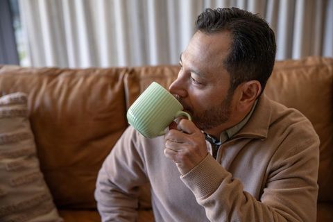 Mature asian man sipping coffee on leather sofa wearing cozy quarter-zip sweater at home