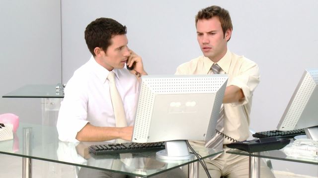 Two businessmen are collaborating at a computer in a modern office setting. They appear to be engaged in a productive discussion, possibly planning or problem-solving. This video is ideal for depicting themes of teamwork, professionalism, corporate environments, and the use of technology in business settings.