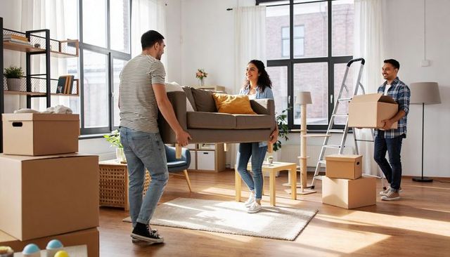 Friends lifting sofa and carrying boxes in bright urban apartment during moving day