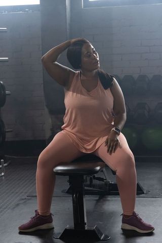 Plus-size african american woman sitting on weight bench wearing pink activewear and smartwatch