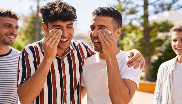Two hispanic friends laughing and wiping eyes with tissues outdoors in sunny courtyard