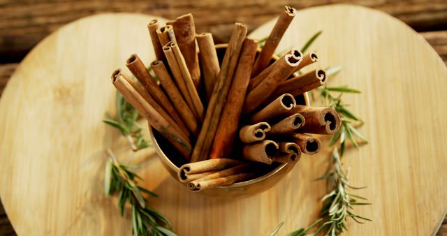 A bunch of cinnamon sticks is presented in a small bowl on a wooden surface, surrounded by sprigs of rosemary, creating a rustic and aromatic display. Cinnamon is often used in cooking and baking for its warm, spicy flavor and is also appreciated for its potential health benefits.
