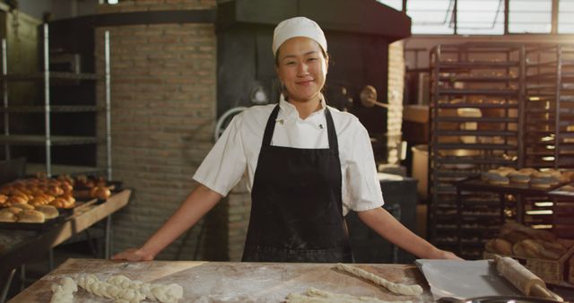 Female baker in professional kitchen smiling confidently at camera. Freshly baked bread placed on tables and shelves. Use for themes related to baking, profession, culinary art, or cooking lifestyle imagery.