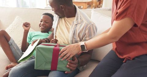 Family exchanging gift and reading card on sofa, parents presenting surprise moment