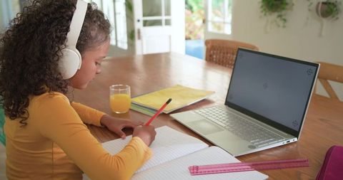 Young girl studying at home wearing wireless headphones writing in notebook with laptop