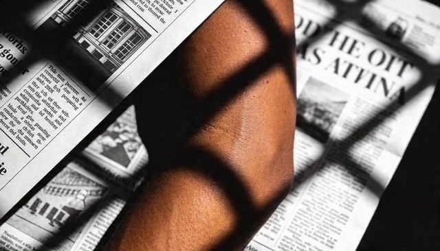 Resting forearm casting window-blind shadows across broadsheet newspaper, high-contrast