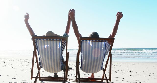 Caucasian senior couple sitting in sunbeds and holding hands in air on beach, copy space. Retirement, leisure, vacations, nature and togetherness, concept, unaltered.