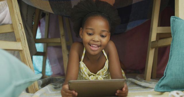 Young African American girl smiling while using tablet inside improvised blanket fort. Perfect for themes related to childhood, technology in education, leisure activities for kids, or imaginative play.