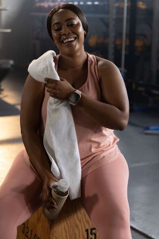 African-american woman resting on plyo box in pink activewear holding towel and shaker