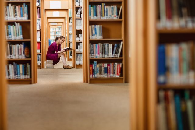 Young woman sitting on floor between bookshelves, deeply engaged in reading a book. Ideal for use in educational materials, college brochures, library promotions, and articles about learning and academic life.