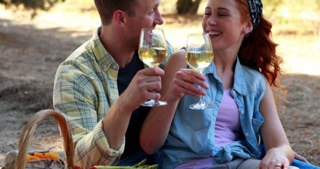 A Caucasian couple enjoys a romantic picnic outdoors, toasting with glasses of white wine, with copy space. Their cheerful interaction and casual attire suggest a relaxed and intimate moment in a natural setting.