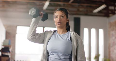 Young woman performing overhead dumbbell press in sunlit industrial fitness studio