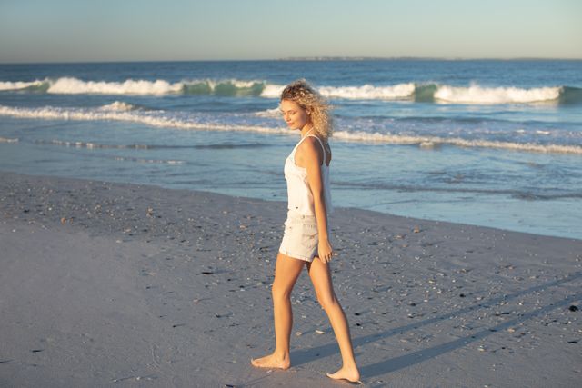 Blonde woman strolling along sandy beach, enjoying peaceful coastal environment during sunset. Ideal for beach vacations, travel blogs, wellness and relaxation content, summer promotions, lifestyle imagery.