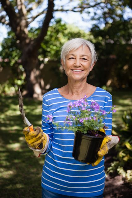 Portrait of smiling senior woman holding potted plant and trowel at backyard