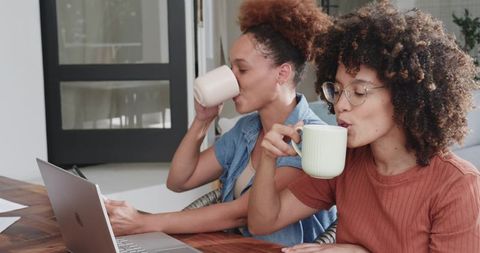 African American Women Sipping Coffee and Collaborating on Laptop in Bright Home Workspace