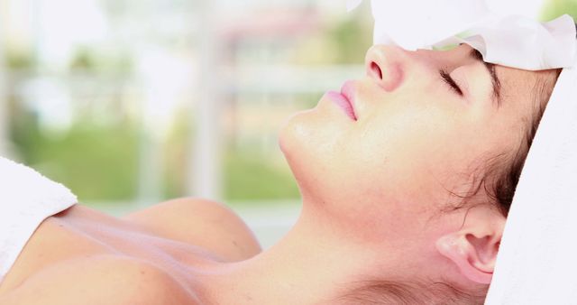Woman resting with eyes closed, enjoying a facial treatment with white towel on her forehead. Perfect for use in content related to wellness, self-care, beauty treatments, skincare regimes, and spa services promotion.