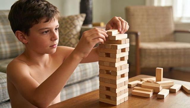 Shirtless boy concentrating while stacking wooden blocks tower in cozy living room