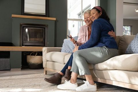 Senior female partners sitting on sofa sharing remote and smiling in cozy living room
