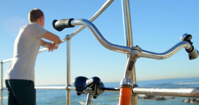 Young man leaning on a railing by the ocean, with bicycle handlebars and a clear blue sky. Ideal for themes of relaxation, outdoor activities, leisure, and summer vacations. Useful in travel brochures, lifestyle blogs, and cycling community publications.