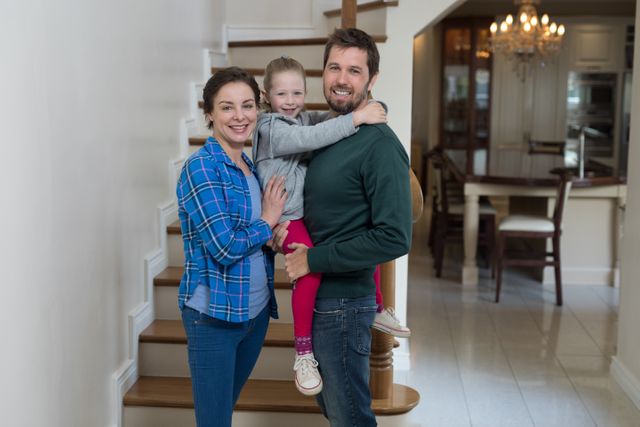 Family of three standing on staircase in modern home, smiling and hugging. Ideal for use in family-oriented advertisements, lifestyle blogs, home decor promotions, and articles about family bonding and happiness.