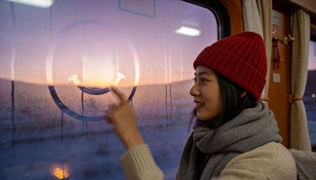 Traveler tracing smiley on fogged train window at sunrise, cozy beanie and scarf, warm mood