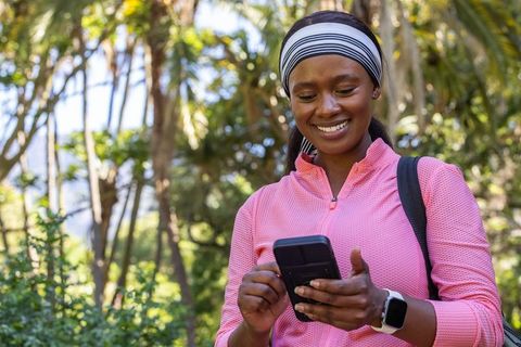 African american woman walking forest trail, smiling and using phone for outdoor fitness