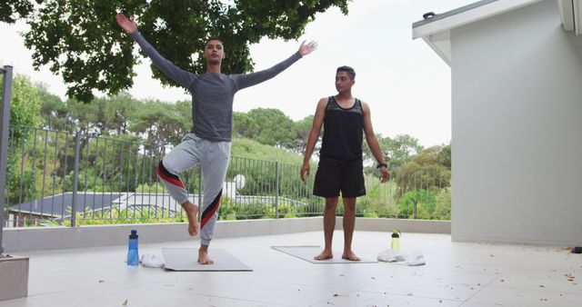 Two young men practicing yoga on an outdoor patio, one performing a tree pose while the other watches. Both are on yoga mats. Diverse use scenarios include wellness blogs, fitness platforms, yoga class promotions, and lifestyle articles focusing on outdoor activities.