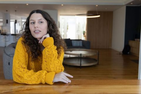 Thoughtful woman leaning on wooden countertop wearing mustard sweater in open-plan living room