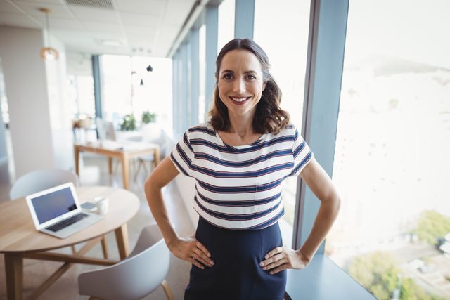 Professional businesswoman standing confidently with hands on hips and smiling in a modern office setting. This can be used for portraying confidence, leadership, and a positive work environment. Suitable for corporate websites, business presentations, promotional materials, and career-related content.