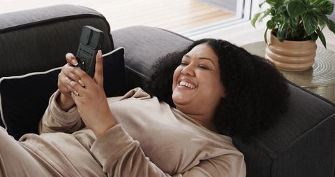 Laughing woman reclining on sofa holding smartphone in cozy loungewear relaxing at home