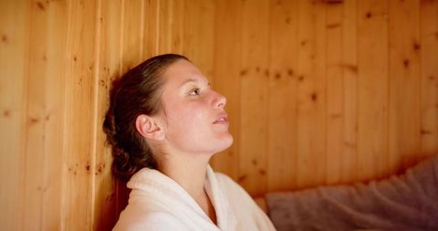 Young woman relaxing in wooden sauna wearing white robe during spa wellness retreat