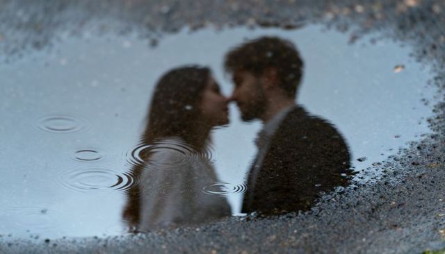 Romantic couple kissing reflected in rain puddle with ripples on urban pavement