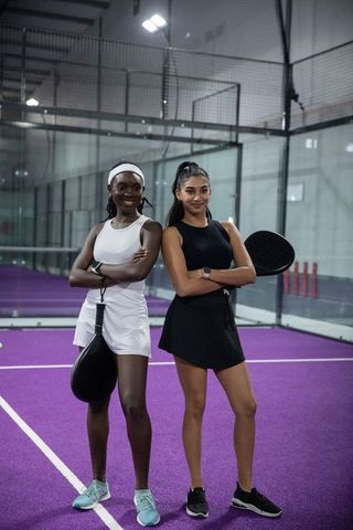 Diverse female padel partners posing on purple indoor court holding paddles and smiling confidently