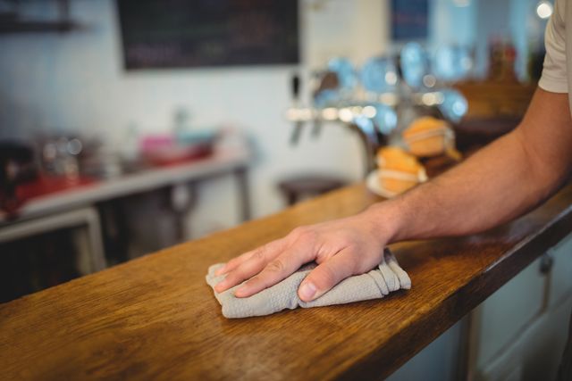Waiter wiping down a wooden counter with a cloth in a cafe. Ideal for use in articles or advertisements related to restaurant hygiene, cleanliness, and maintenance. Can also be used in hospitality industry training materials or service-oriented promotions.