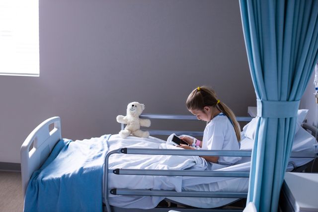 Young girl sitting in a hospital bed, engaging with a digital tablet, with a teddy bear beside her. Suitable for use in healthcare-related marketing, pediatric medical blogs, patient care promotions, and educational materials on technology in healthcare.