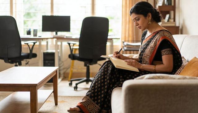 Indian woman writing notes on sofa with home workstation and dual monitors for remote work
