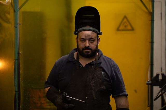 Caucasian male factory worker wearing welding mask, standing in industrial setting, looking at camera. Ideal for use in articles about manufacturing, industrial safety, skilled labor, and engineering. Can be used in promotional materials for industrial equipment, safety gear, and workforce training programs.