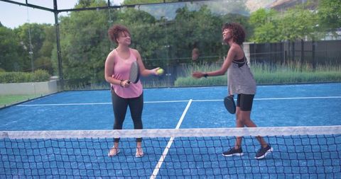 African american women playing paddle sport on blue court, laughing and sharing ball