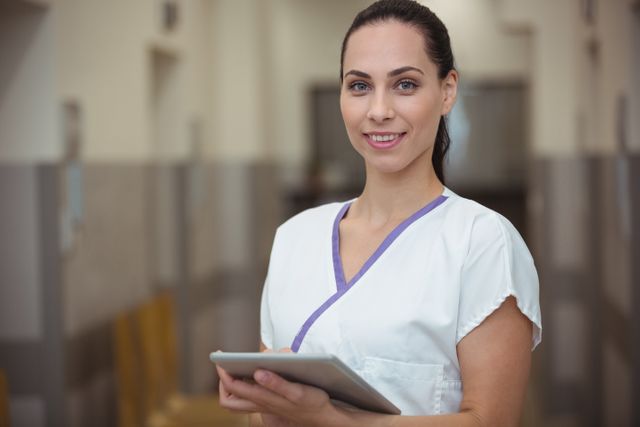 This image depicts a smiling female nurse holding and using a digital tablet in a hospital corridor. Ideal for use in healthcare-related content, medical websites, hospital brochures, and articles about modern healthcare technology and patient care.