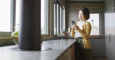 Asian woman using smartphone in modern minimalist kitchen by window, healthy lifestyle